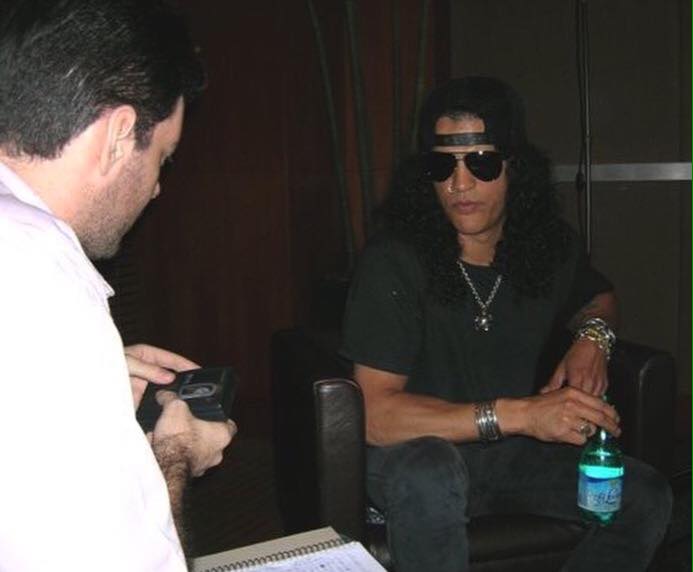 Foto dos bastidores de entrevista com o guitarrista Slash. No lado esquerdo aparece Henrique Inglez de Souza, preparando o gravador, e no lado direito, Slash. O guitarrista está de boné e óculos escuros.